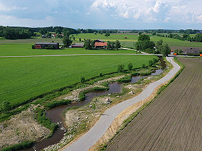 Ein Geh- und Radweg verläuft parallel zu einem renaturierten Bach mit geschwungenem Verlauf durch eine landwirtschaftlich geprägte Landschaft. Ein Geh- und Radweg verläuft parallel zu einem renaturierten Bach mit geschwungenem Verlauf durch eine landwirtschaftlich geprägte Landschaft.