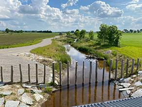 Blick von einem Drosselbauwerk auf einen Bach und einen Grobrechen aus großen Holzstämmen. Im Hintergrund verläuft der Bach leicht geschwungen durch die Landschaft. Ein breiter Kiesweg verläuft links parallel dazu. Rechts schließt sich eine Baumreihe und Grünland an. Blick von einem Drosselbauwerk auf einen Bach und einen Grobrechen aus großen Holzstämmen. Im Hintergrund verläuft der Bach leicht geschwungen durch die Landschaft. Ein breiter Kiesweg verläuft links parallel dazu. Rechts schließt sich eine Baumreihe und Grünland an.