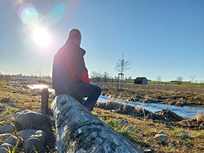 Ein Mann sitzt auf einem entrindeten Baumstamm am Ufer eines Baches. Die Sonne spiegelt sich im Wasser des leicht geschwungen fließenden Baches. Im Hintergrund landwirtschaftliche Flächen und eine Hütte. Ein Mann sitzt auf einem entrindeten Baumstamm am Ufer eines Baches. Die Sonne spiegelt sich im Wasser des leicht geschwungen fließenden Baches. Im Hintergrund landwirtschaftliche Flächen und eine Hütte.