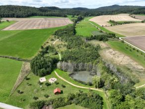 Luftaufnahme einer Grünfläche mit Spielplatz und Hütte sowie einem Teich. Über den Zulauf zum Bach führt ein Weg mit Trittsteinen. Es schließt sich eine Auenfläche mit Wald und Offenland an, durch die ein Weg hindurchführt. Im Hintergrund Acker- und Grünlandflächen, dahinter bewaldete Hügel.