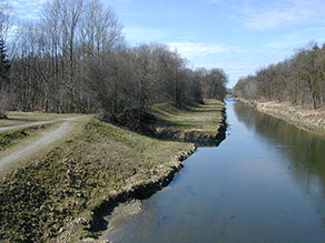 Blick auf einen Fluss mit begradigten Ufern und ohne Breitenvarianz. Ein Hochwasserschutzdeich verläuft parallel zum Gewässer mit wenig Abstand zum Ufer. Die Fläche zwischen Deich und Fluss ist grasbewachsen und von Gehölzen weitestgehend freigehalten. Hinter dem Deich erstreckt sich ein Waldgebiet. Blick auf einen Fluss mit begradigten Ufern und ohne Breitenvarianz. Ein Hochwasserschutzdeich verläuft parallel zum Gewässer mit wenig Abstand zum Ufer. Die Fläche zwischen Deich und Fluss ist grasbewachsen und von Gehölzen weitestgehend freigehalten. Hinter dem Deich erstreckt sich ein Waldgebiet.