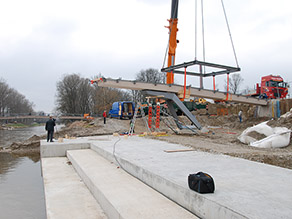 Bauarbeiten an einem neuen Aussichtssteg am Ufer eines Flusses: Ein Kran hebt den Steg ein, mehrere Personen sind mit der Montage beschäftigt. Im Vordergrund führen mehrere Treppenstufen aus Beton ans Wasser. Bauarbeiten an einem neuen Aussichtssteg am Ufer eines Flusses: Ein Kran hebt den Steg ein, mehrere Personen sind mit der Montage beschäftigt. Im Vordergrund führen mehrere Treppenstufen aus Beton ans Wasser.