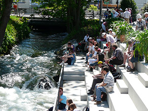 Am Zusammenfluss des Mühlbachs und des Hammerbachs am Ichikawaplatz strömt Wasser über Felsen und zahlreiche Personen sitzen daneben durch ein Geländer getrennt auf Sitzstufen in der Sonne. Am Zusammenfluss des Mühlbachs und des Hammerbachs am Ichikawaplatz strömt Wasser über Felsen und zahlreiche Personen sitzen daneben durch ein Geländer getrennt auf Sitzstufen in der Sonne.