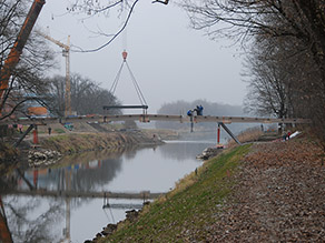 Über einen Fluss wird das Element einer neuen Fußgängerbrücke eingehoben. Mehrere Personen sind mit der Montage auf der Brücke beschäftigt. Über einen Fluss wird das Element einer neuen Fußgängerbrücke eingehoben. Mehrere Personen sind mit der Montage auf der Brücke beschäftigt.