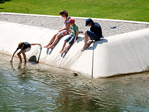 Auf einem zum Wasser abfallenden Gestaltungselement aus Beton an einem Bachufer sitzen mehrere Personen. Ein Kind steigt in das flache Wasser im Randbereich. Auf einem zum Wasser abfallenden Gestaltungselement aus Beton an einem Bachufer sitzen mehrere Personen. Ein Kind steigt in das flache Wasser im Randbereich.