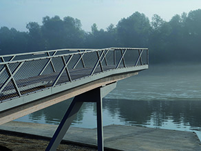 Ein Aussichtssteg mit Geländer ragt über das betonierte Ufer eines Flusses, dessen gegenüberliegendes Ufer im Hintergrund im Nebel verschwindet. Ein Aussichtssteg mit Geländer ragt über das betonierte Ufer eines Flusses, dessen gegenüberliegendes Ufer im Hintergrund im Nebel verschwindet.