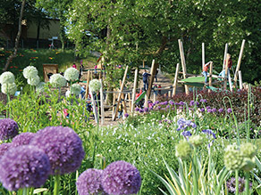 Blick über blühende Blumenbeete hinweg auf einen Spielplatz mit Holzklettergerüst. Mehrere Kinder klettern auf dem Gerüst. Blick über blühende Blumenbeete hinweg auf einen Spielplatz mit Holzklettergerüst. Mehrere Kinder klettern auf dem Gerüst.