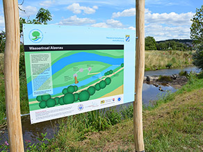 Eine Infotafel mit Holzpfosten steht am Ufer eines Flusses und informiert über die Wasserinsel Alzenau. Eine Infotafel mit Holzpfosten steht am Ufer eines Flusses und informiert über die Wasserinsel Alzenau.