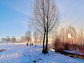 Drei Personen spazieren auf einem Weg parallel zu einem Bach durch eine winterliche Schneelandschaft bei Sonnenuntergang. Drei Personen spazieren auf einem Weg parallel zu einem Bach durch eine winterliche Schneelandschaft bei Sonnenuntergang.