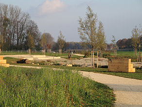 Ein Geh- und Radweg verläuft geschwungen und parallel zu einem Bach durch eine Grünanlage. Zwei Holzpodeste stehen am Wegesrand. Die abgeflachten Kiesufer des Bachs sind weitestgehend unbewachsen. Im Hintergrund zwei Sitzreihen aus Natursteinquadern in der Böschung. Ein Geh- und Radweg verläuft geschwungen und parallel zu einem Bach durch eine Grünanlage. Zwei Holzpodeste stehen am Wegesrand. Die abgeflachten Kiesufer des Bachs sind weitestgehend unbewachsen. Im Hintergrund zwei Sitzreihen aus Natursteinquadern in der Böschung.