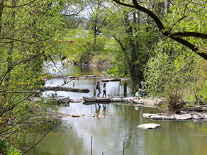 Ein schmaler Übergang aus Trittsteinen führt über einen Fluss von einem zum anderen Ufer. Mehrere Personen stehen auf den Steinen. Die Ufer sind mit Gehölzen dicht bewachsen. Ein schmaler Übergang aus Trittsteinen führt über einen Fluss von einem zum anderen Ufer. Mehrere Personen stehen auf den Steinen. Die Ufer sind mit Gehölzen dicht bewachsen.