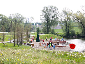 Am Flachufer eines Flusses spielen zahlreiche Kinder an einem Wasserspielplatz. Bänke stehen auf einer Wiesenfläche daneben. Am Flachufer eines Flusses spielen zahlreiche Kinder an einem Wasserspielplatz. Bänke stehen auf einer Wiesenfläche daneben.