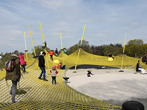 Auf einem Spielplatz mit Sandfläche und einem Gerüst aus Netzen spielen zahlreiche Kinder und Erwachsene. Auf einem Spielplatz mit Sandfläche und einem Gerüst aus Netzen spielen zahlreiche Kinder und Erwachsene.