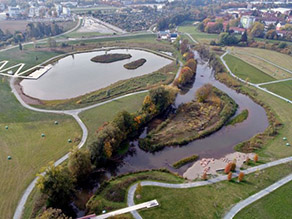 Luftaufnahme des Roten Mains mit zwei Gewässerarmen, die eine Insel bilden. Im Vordergrund ein Wasserspielplatz mit Sandstrand und Flachwasserbereich am Flussufer. Mehrere Wege führen entlang des Gewässers. Wiesenflächen und ein großer Landschaftssee mit Seebühne und zwei Inseln prägen die angrenzende Grünanlage. Luftaufnahme des Roten Mains mit zwei Gewässerarmen, die eine Insel bilden. Im Vordergrund ein Wasserspielplatz mit Sandstrand und Flachwasserbereich am Flussufer. Mehrere Wege führen entlang des Gewässers. Wiesenflächen und ein großer Landschaftssee mit Seebühne und zwei Inseln prägen die angrenzende Grünanlage.