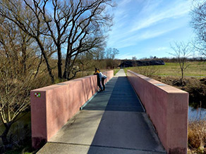 Auf einer Brücke über einen Fluss mit Betonmauern als Brüstung stehen zwei Personen und betrachten das Gewässer. Auf einer Brücke über einen Fluss mit Betonmauern als Brüstung stehen zwei Personen und betrachten das Gewässer.
