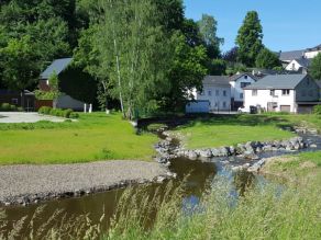 Ein Fluss mit sehr flachen Ufern und daran angrenzenden Kies- und Wiesenflächen teilt sich in zwei Gewässerarme auf. Im Hintergrund Teil eines Wohnmobilstellplatzes und Wohnbebauung. Ein Fluss mit sehr flachen Ufern und daran angrenzenden Kies- und Wiesenflächen teilt sich in zwei Gewässerarme auf. Im Hintergrund Teil eines Wohnmobilstellplatzes und Wohnbebauung.