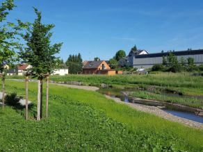 Ein Fluss mit flachem Kiesufer und daran angrenzenden Wiesenflächen. Im Hintergrund Lagerhallen und Wohnbebauung. Ein Fluss mit flachem Kiesufer und daran angrenzenden Wiesenflächen. Im Hintergrund Lagerhallen und Wohnbebauung.