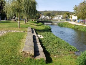 Am abgeflachten Ufer eines Flusses zwei Sitzreihen aus Steinquadern. Weiden wachsen entlang des Ufers. Im Hintergrund eine Fußgängerbrücke über den Fluss. Am abgeflachten Ufer eines Flusses zwei Sitzreihen aus Steinquadern. Weiden wachsen entlang des Ufers. Im Hintergrund eine Fußgängerbrücke über den Fluss.