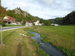 Der neue geschwungene Bachlauf weist flache, mit Gras bewachsene Ufer auf. Eine kurze Treppe führt ans Wasser. Rechts vom Bach schließt sich Grünland an. Links vom Bach eine Straße. Im Hintergrund mehrere Häuser und ein Hügel mit Felsen und einer Ruine.