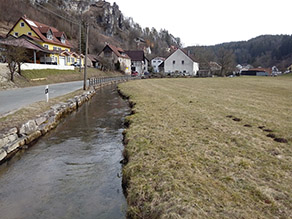 Ein Bach fließt unmittelbar neben einer Straße. Das Ufer zur Straße hin ist mit einer Natursteinmauer geschützt. Im Hintergrund verläuft zusätzlich ein Geländer zwischen Straße und Bach. Auf der anderen Bachseite schließt sich Grünland an. Im Hintergrund mehrere Häuser und ein Hügel mit Felsen und einer Ruine.