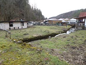Ein Bach fließt durch einen Ort. Die Ufer sind mit Steinmauern verbaut. Im Hintergrund ein Gewerbehalle und mehrere Schuppen.