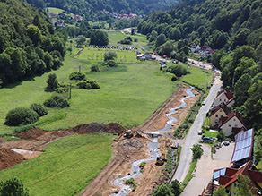 Tal der Trubach von oben während der Baumaßnahme: Der alte Bachlauf verläuft unmittelbar neben einer Straße. Daneben der neue, geschwungene Gewässerlauf mit abgeflachten Ufern. Ein Rahmendurchlass ermöglicht die Überfahrt  über den neuen Gewässerlauf. Zwei Bagger stehen am Ufer. Es schließt sich Grünland an. Im Hintergrund ein Fußballfeld.