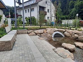 Ein Bach fließt aus einem Durchlass. Das Wasser ist sehr klar, die Gewässersohle besteht aus Kies. Mehrere Trittsteine im Bach ermöglichen die Querung. Gepflasterte Treppenstufen führen zum Bach. Am Rand der gepflasterten Fläche Sitzsteine und eine Bank.