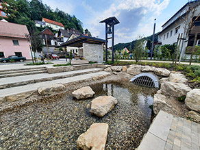 Im Vordergrund fließt der Bach aus einem Durchlass. Das Wasser ist sehr klar, die Gewässersohle besteht aus Kies. Mehrere Trittsteine im Bach ermöglichen die Querung. Am gegenüberliegenden Ufer führen mehrere Treppenstufen an das Wasser. Eine Bank sowie Steinquader mit Holzauflage laden zum Verweilen ein. Im Hintergrund ein überdachter Rastplatz bestehend aus zwei Natursteinmauern und einem Dachgerüst aus Stahl und Holz sowie ein Glockengerüst. Dahinter eine Siedlung.