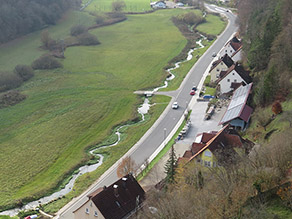 Tal der Trubach von oben: Der neue, geschwungene Gewässerlauf verläuft neben einer Straße. Eine kleine Brücke führt über den Bach. Der verbleibende Talraum besteht überwiegend aus Grünland und kleineren Gehölzen.