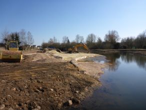 Ein Bagger und eine Walze stehen am abgeflachten Ufer eines Flusses. Kies und Erdhaufen daneben. Eine halbkreisförmige Mauer aus großformatigen Natursteinen mit vorgelagerter Kiesfläche entsteht am Ufer. Ein Bagger und eine Walze stehen am abgeflachten Ufer eines Flusses. Kies und Erdhaufen daneben. Eine halbkreisförmige Mauer aus großformatigen Natursteinen mit vorgelagerter Kiesfläche entsteht am Ufer.