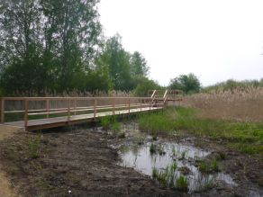 Ein Holzsteg mit Geländer führt über eine teilweise mit Wasser bestandene Fläche in ein Röhricht. Am Ende des Steges eine erhöhte Aussichtsplattform. Ein Holzsteg mit Geländer führt über eine teilweise mit Wasser bestandene Fläche in ein Röhricht. Am Ende des Steges eine erhöhte Aussichtsplattform.