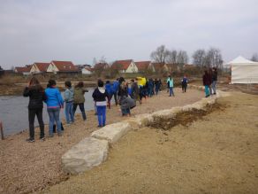 Eine Schulklasse steht auf einem Kiesstrand am Ufer eines Gewässers. Die flach zum Wasser abfallende Kiesfläche ist durch eine geschwungene, niedrige Natursteinmauer begrenzt. Dahinter schließt sich eine noch brach liegende Fläche mit einem Pavillon an. Eine Schulklasse steht auf einem Kiesstrand am Ufer eines Gewässers. Die flach zum Wasser abfallende Kiesfläche ist durch eine geschwungene, niedrige Natursteinmauer begrenzt. Dahinter schließt sich eine noch brach liegende Fläche mit einem Pavillon an.