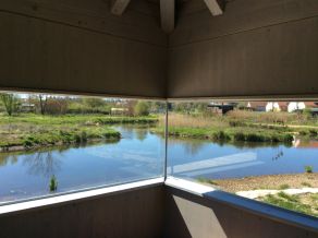 Blick aus dem Innern eines Pavillons durch ein Sichtfenster auf eine Gewässerlandschaft. Im Hintergrund eine grasbewachsene Insel, die von Wasser umgeben ist. Blick aus dem Innern eines Pavillons durch ein Sichtfenster auf eine Gewässerlandschaft. Im Hintergrund eine grasbewachsene Insel, die von Wasser umgeben ist.