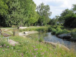Ein aufgeweiteter Bach mit Fachwasserbereich, abgeflachten Ufern und einem Treppenabgang. Eine bunte Blühwiese bildet die flache Uferböschung im Vordergrund. Eine Holzbank und ein Steinquader bieten Sitzmöglichkeiten am Bachufer mit Blick auf das Gewässer. Parallel zum Bach verläuft ein asphaltierter Geh- und Radweg. Ein aufgeweiteter Bach mit Fachwasserbereich, abgeflachten Ufern und einem Treppenabgang. Eine bunte Blühwiese bildet die flache Uferböschung im Vordergrund. Eine Holzbank und ein Steinquader bieten Sitzmöglichkeiten am Bachufer mit Blick auf das Gewässer. Parallel zum Bach verläuft ein asphaltierter Geh- und Radweg.