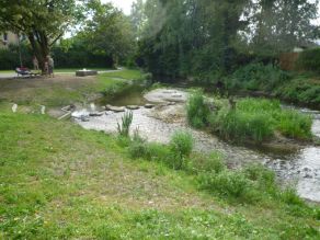 Erlebnisbereich an einem verzweigten Bach mit Treppenstufen an das Wasser und Trittsteinen über einen Gewässerarm zu einer teilweise mit Gras bewachsenen Kiesinsel. Der kiesige Bachlauf ist flach überströmt. Im Hintergrund befinden sich einige Personen an einer Holzliege und einem großen Steinquader am Rand eines Geh- und Radweges, der entlang des Gewässers verläuft. Erlebnisbereich an einem verzweigten Bach mit Treppenstufen an das Wasser und Trittsteinen über einen Gewässerarm zu einer teilweise mit Gras bewachsenen Kiesinsel. Der kiesige Bachlauf ist flach überströmt. Im Hintergrund befinden sich einige Personen an einer Holzliege und einem großen Steinquader am Rand eines Geh- und Radweges, der entlang des Gewässers verläuft.