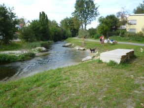 Leicht geschwungener Bach mit Gewässeraufweitung und flach abfallenden Ufern. Ein Treppenabgang ermöglicht Zugang zum Bach. Zwei Steinquader und mehrere Findlinge laden als Sitzgelegenheiten ein. Drei Personen und ein Hund stehen am grasbewachsenen Ufer. Ein Geh- und Radweg verläuft entlang des Gewässers. Leicht geschwungener Bach mit Gewässeraufweitung und flach abfallenden Ufern. Ein Treppenabgang ermöglicht Zugang zum Bach. Zwei Steinquader und mehrere Findlinge laden als Sitzgelegenheiten ein. Drei Personen und ein Hund stehen am grasbewachsenen Ufer. Ein Geh- und Radweg verläuft entlang des Gewässers.