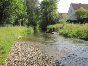 Ein flachüberströmter, kiesiger Bachlauf wird im Vordergrund von einer Kiesfläche und im Hintergrund von hohem Gras und Bäumen gesäumt. Ein Kind steht im Wasser, ein Mann sitzt auf einer Holzbuhne daneben. Ein flachüberströmter, kiesiger Bachlauf wird im Vordergrund von einer Kiesfläche und im Hintergrund von hohem Gras und Bäumen gesäumt. Ein Kind steht im Wasser, ein Mann sitzt auf einer Holzbuhne daneben.