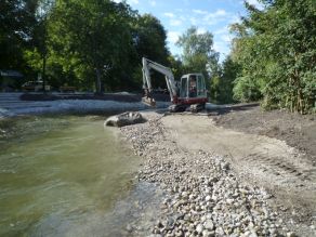 Ein kleiner Bagger führt Modellierungsarbeiten am neuen, kiesigen Flachufer eines Bachs durch. Im Hintergrund führt ein Treppenabgang zum Wasser. Ein kleiner Bagger führt Modellierungsarbeiten am neuen, kiesigen Flachufer eines Bachs durch. Im Hintergrund führt ein Treppenabgang zum Wasser.