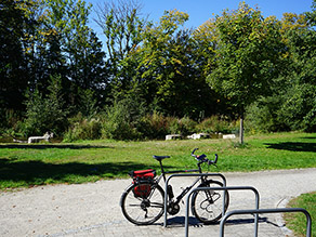 Ein Fahrrad steht an einem Fahrradständer aus Metall auf einer gepflasterten Fläche neben einem Kiesweg. Im Hintergrund sind eine Wiesenfläche, Bäume und Sträucher am Ufer eines Gewässers zu sehen. Ein Fahrrad steht an einem Fahrradständer aus Metall auf einer gepflasterten Fläche neben einem Kiesweg. Im Hintergrund sind eine Wiesenfläche, Bäume und Sträucher am Ufer eines Gewässers zu sehen.