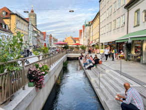 Durch den Stadtplatz an der Oberen Hauptstraße in Freising fließt der freigelegte Moosach-Kanal. Stufen führen vom Stadtplatzniveau hinab zum Wasser und bieten auch Sitzgelegenheiten, auf denen Menschen mit Eiscreme sitzen. Die gegenüberliegende Kanalseite ist mit Kübelpflanzen begrünt. Durch den Stadtplatz an der Oberen Hauptstraße in Freising fließt der freigelegte Moosach-Kanal. Stufen führen vom Stadtplatzniveau hinab zum Wasser und bieten auch Sitzgelegenheiten, auf denen Menschen mit Eiscreme sitzen. Die gegenüberliegende Kanalseite ist mit Kübelpflanzen begrünt.