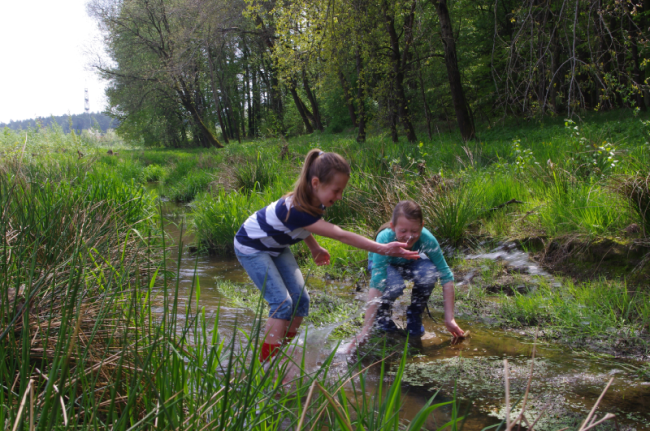 Zwei Kinder stehen mit Gummistiefeln in einem flachen Bach und spritzen mit Wasser um sich. Im Hintergrund windet sich der Bach durch hohes Gras.