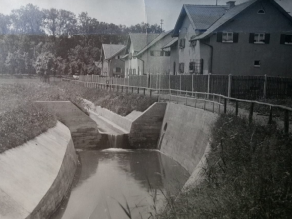 Historische Aufnahme eines stark verbauten Bachs mit einem Absturz und einem begradigten Trapezprofil aus Beton. Neben dem Gewässer ein Gehweg und einige Häuser.