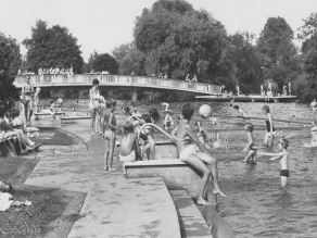 Historische Aufnahme eines Freizeitbads an einem Fluss. Treppen führen ins Wasser und in einen vom restlichen Fluss durch einen Zaun abgetrennten Schwimmbereich. Viele Kinder und Erwachsene spielen und planschen im und am Wasser.