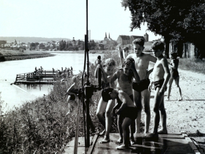 Historische Aufnahme von Kindern, die sich am Ufer eines Flusses unter einer Freilanddusche abduschen. Im Hintergrund ein Holzfloß mit Stegen auf dem Wasser und die Kulisse einer Stadt.