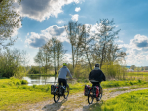 Zwei Fahrradfahrer mit Gepäcktaschen auf einem Schotterweg am Ufer eines Flusses.