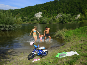 Eine Familie mit Kleinkind kommt in Badekleidung aus dem Wasser eines Flusses im Bereich einer flachen Badebucht. Am Ufer ein Kinderfahrrad und ein Handtuch. Am gegenüberliegenden Ufer ragen Kalkfelsen aus der bewaldeten Talböschung.
