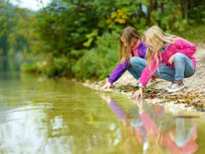 Zwei Mädchen hocken am Kiesufer eines Gewässers und strecken ihre Hände ins flache Wasser.