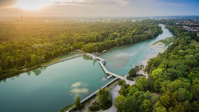 Schrägluftbild eines Flusses mit einer Wehranlage in der Bildmitte und angrenzenden Waldgebieten. Über das Stauwehr verläuft ein Fußgängersteg mit Getriebehäuschen und historischem Glockenturm, der die beiden Flussufer miteinander verbindet. Schrägluftbild eines Flusses mit einer Wehranlage in der Bildmitte und angrenzenden Waldgebieten. Über das Stauwehr verläuft ein Fußgängersteg mit Getriebehäuschen und historischem Glockenturm, der die beiden Flussufer miteinander verbindet.