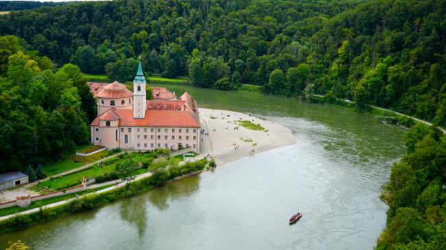 Schrägbildaufnahme eines Klosters mit Glockenturm, das am Ufer eines großen Flusses in dessen Flussschleife und umgeben von dicht bewaldeten Hängen liegt. Vor dem Kloster erstreckt sich eine breite Kiesbank, auf der sich Menschen aufhalten, während ein kleines Boot auf dem Fluss vorbeifährt. Schrägbildaufnahme eines Klosters mit Glockenturm, das am Ufer eines großen Flusses in dessen Flussschleife und umgeben von dicht bewaldeten Hängen liegt. Vor dem Kloster erstreckt sich eine breite Kiesbank, auf der sich Menschen aufhalten, während ein kleines Boot auf dem Fluss vorbeifährt.