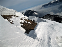 Die mächtige Schneedecke öffnet sich zu einem Gleitschneemaul. Der grasige Boden wird sichtbar. Im Hintergrund sieht man das winterliche Chiemgau mit Nebelresten im Tal.